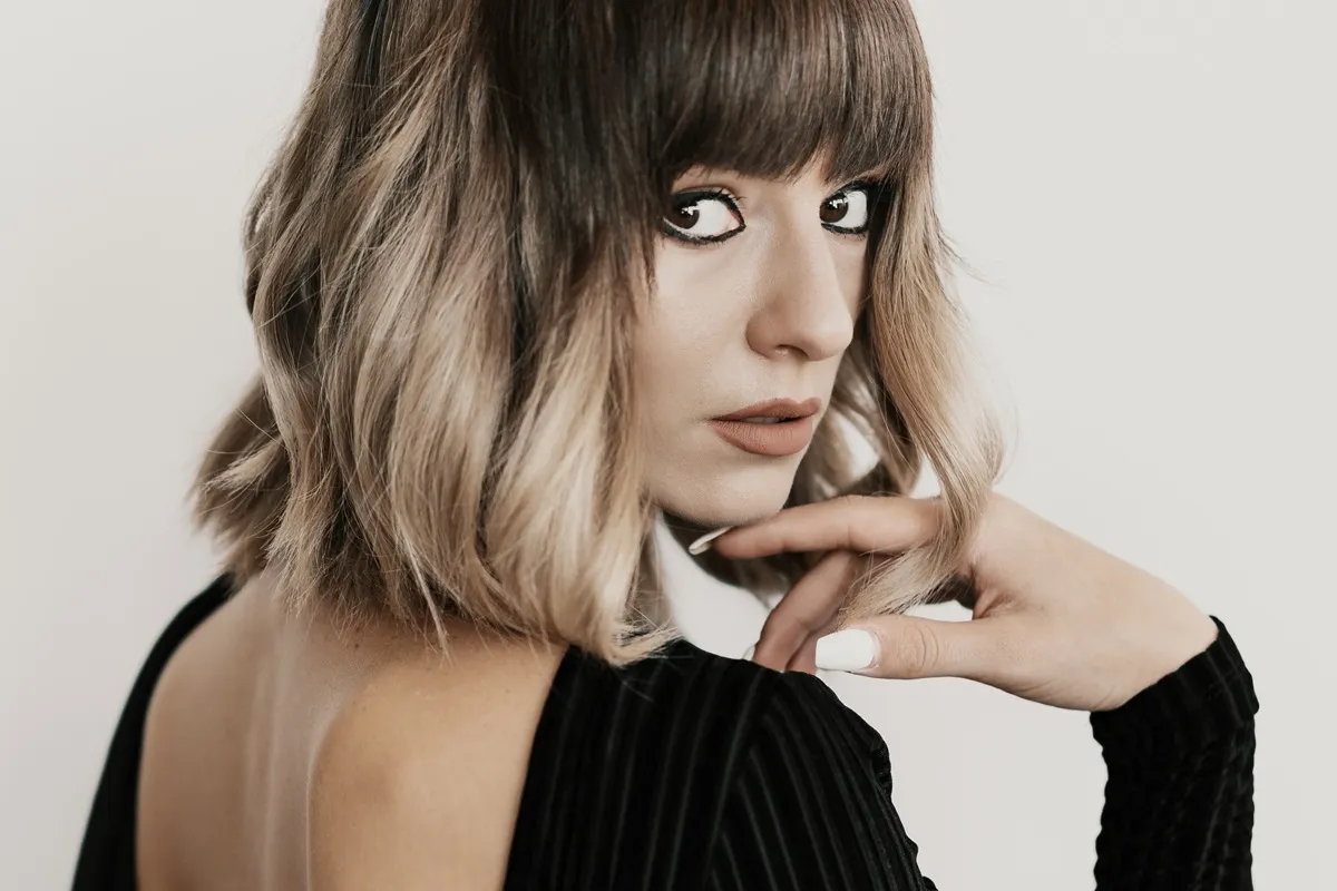 Donna con taglio di capelli medi e frangia piena, capelli biondo cenere con onde leggere, indossa un abito nero con schiena scoperta e guarda verso la fotocamera con espressione intensa.