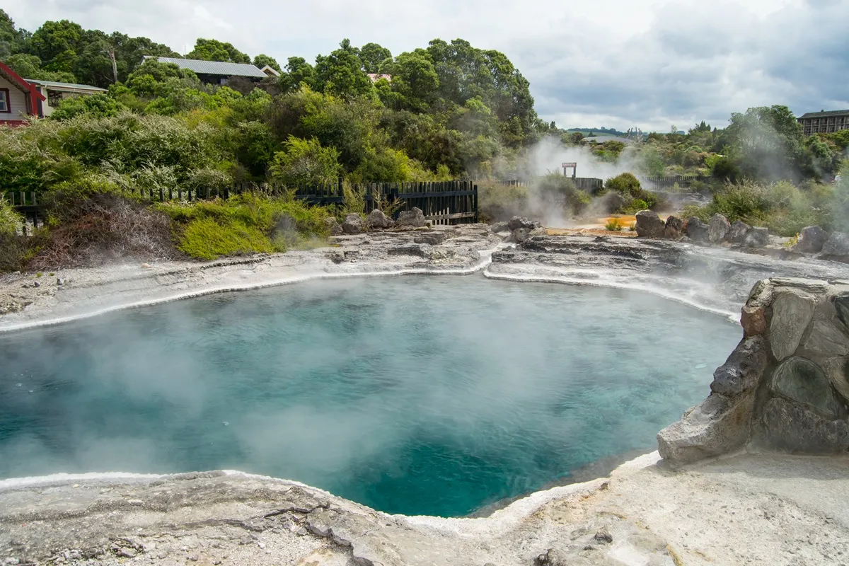 Piscina termale naturale con acqua azzurra e vapori caldi, circondata da rocce e vegetazione verde lussureggiante.