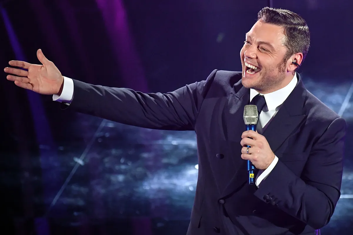 Tiziano Ferro, in abito elegante scuro, canta sorridente con microfono in mano durante una performance sul palco.