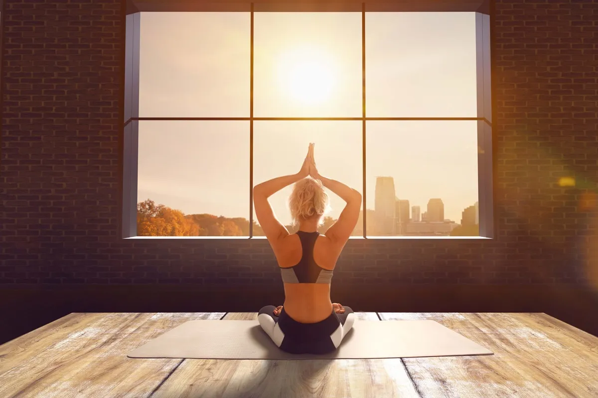 Donna che pratica yoga al mattino in posizione di meditazione davanti a una grande finestra con vista sulla città illuminata dal sole nascente.