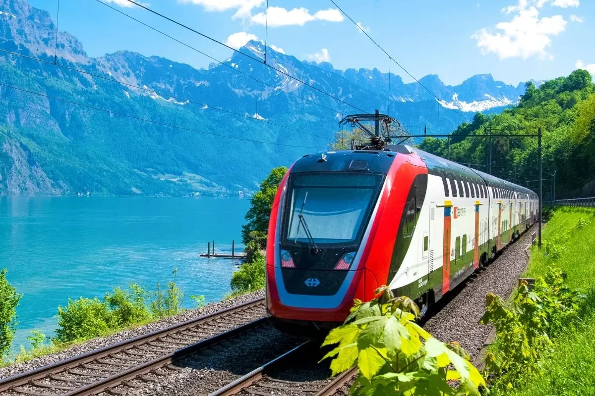Treno rosso e bianco che viaggia lungo un lago alpino con montagne innevate sullo sfondo, simbolo del viaggio Interrail attraverso l’Europa.