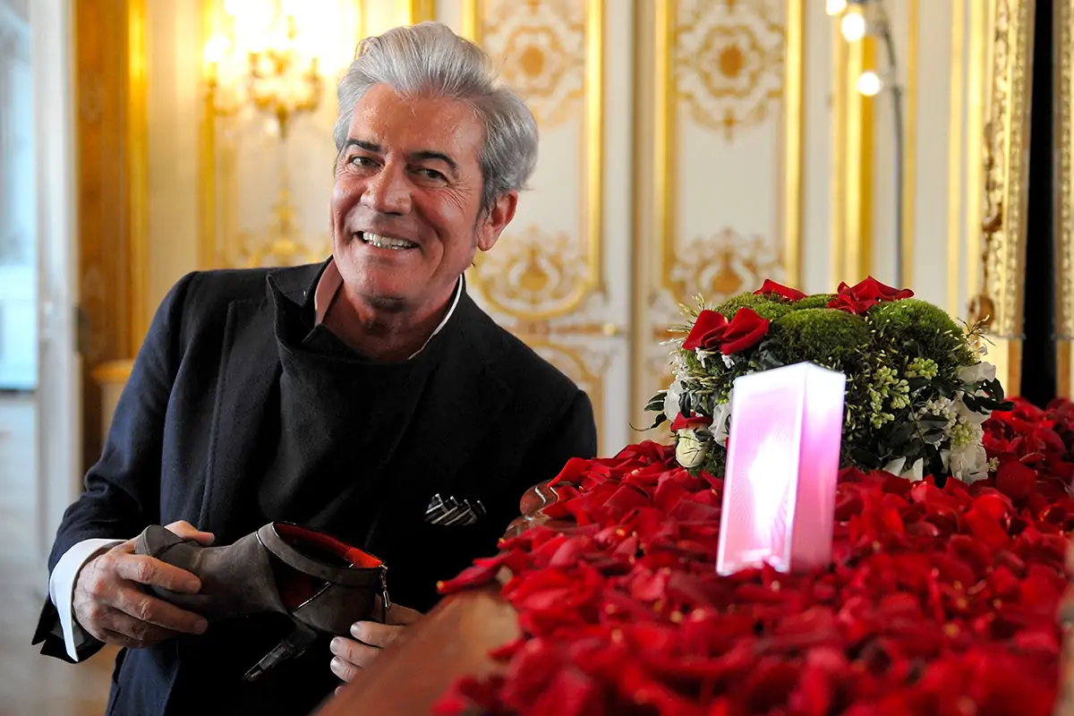 Uomo elegante con capelli grigi e sorriso amichevole, tiene una scarpa di lusso in mano accanto a un tavolo decorato con petali di rosa rossa e fiori, in una sala dorata dallo stile barocco.