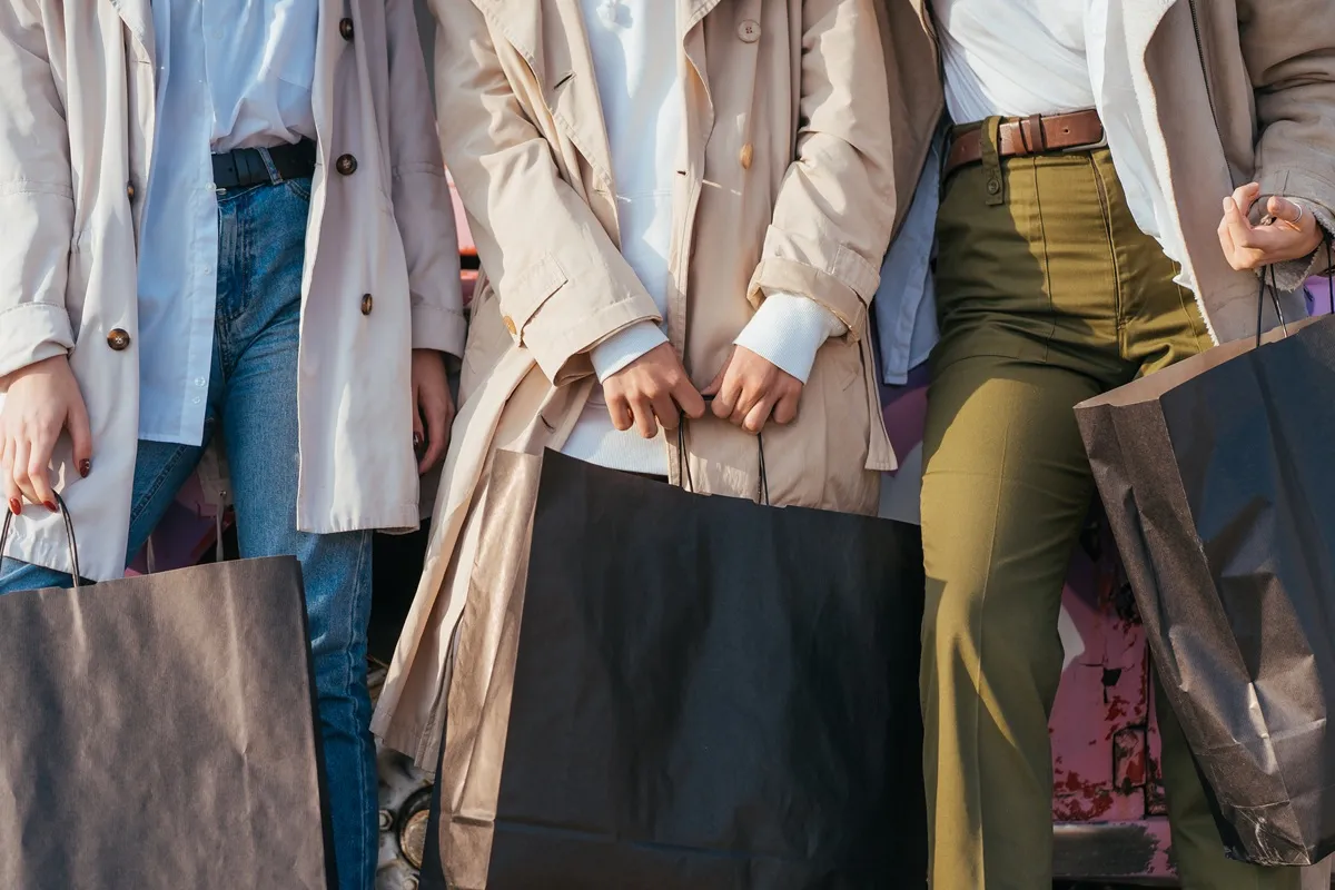 Tre donne vestite con cappotti eleganti e pantaloni alla moda tengono grandi borse da shopping nere durante una giornata di acquisti in città.