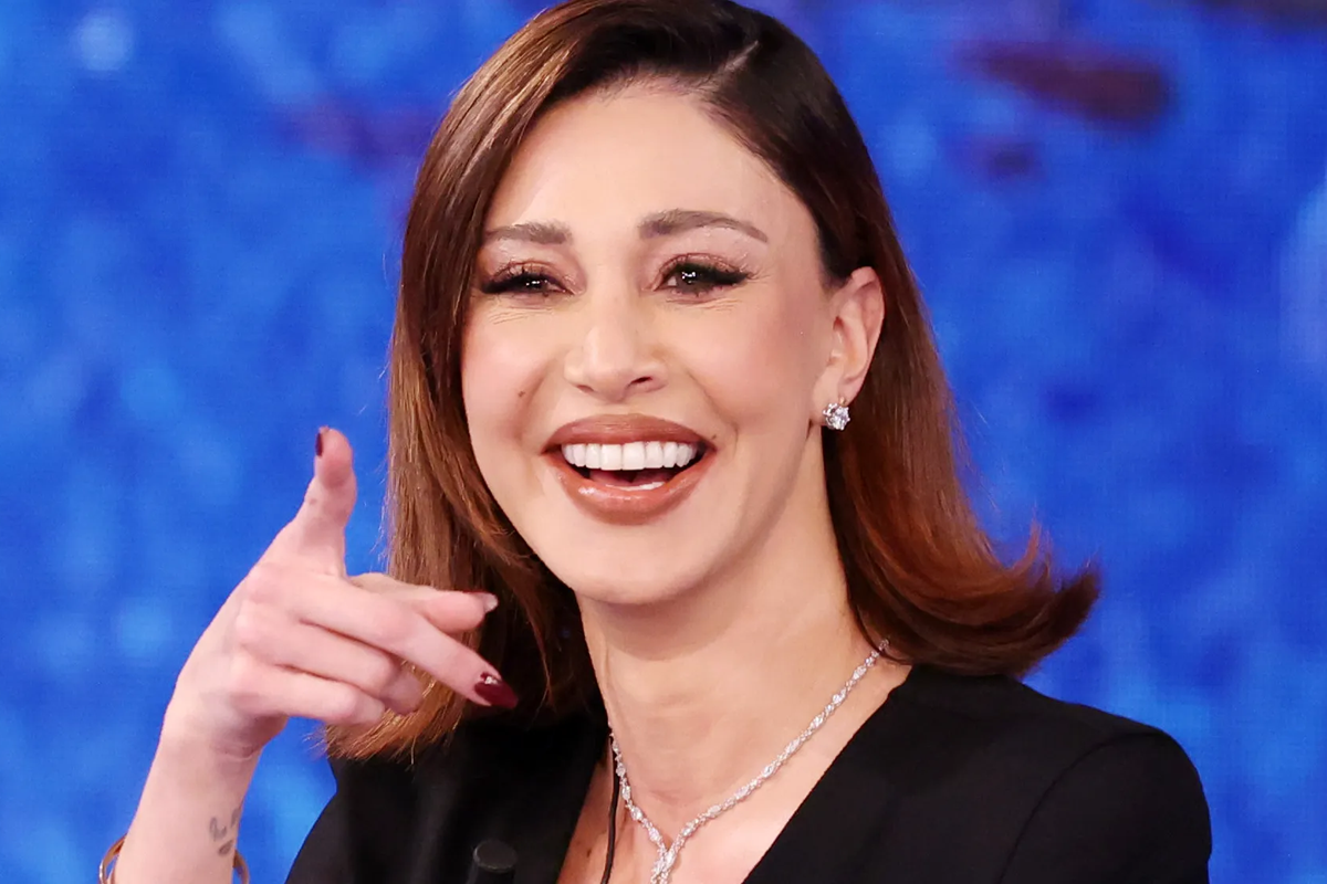 Donna sorridente con capelli castani e trucco naturale, indossa un abito nero e gioielli luminosi mentre gesticola davanti a uno sfondo blu in uno studio televisivo.