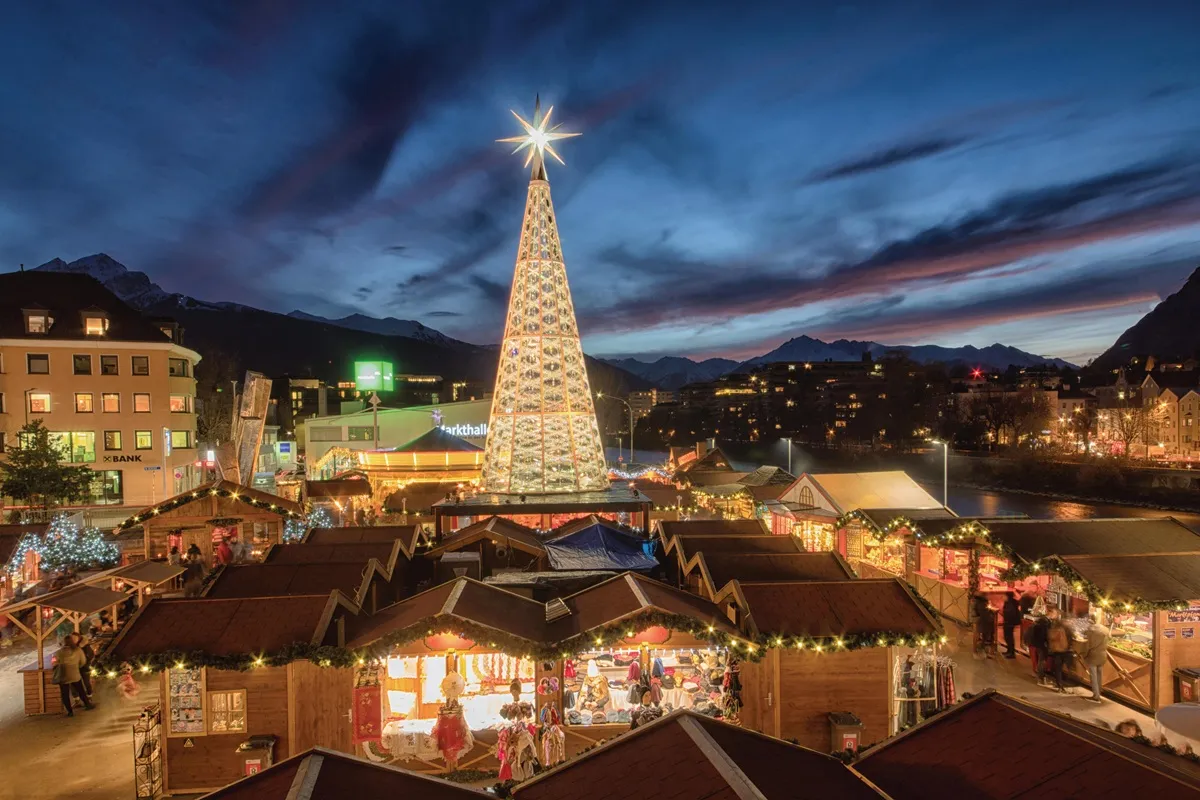 Mercatino di Natale a Innsbruck illuminato al tramonto, con una grande albero di Natale decorato e chalet in legno addobbati, circondati dalle montagne innevate del Tirolo.