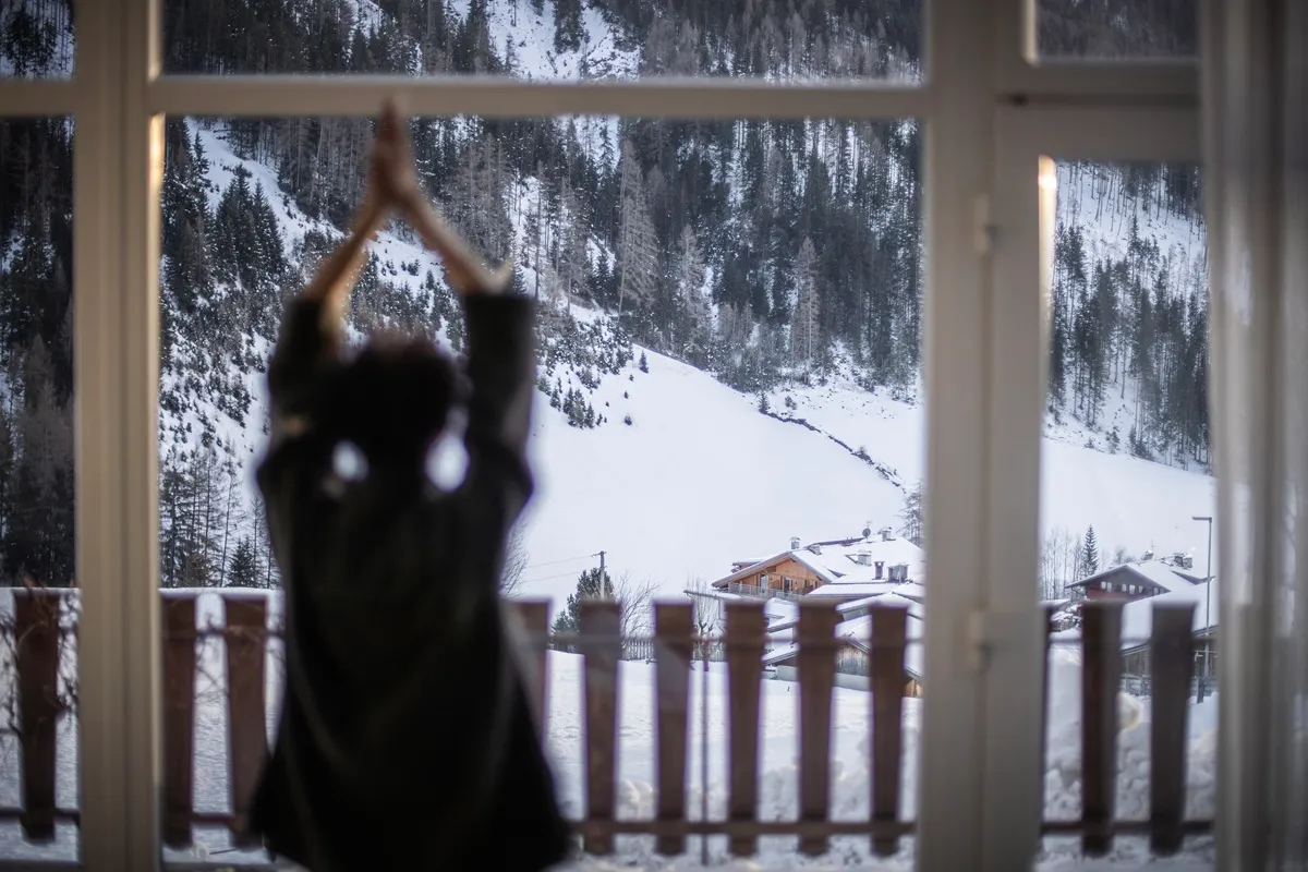 Persona di spalle che fa stretching o yoga davanti a una grande finestra con vista su un paesaggio montano innevato e chalet di legno tra gli alberi.