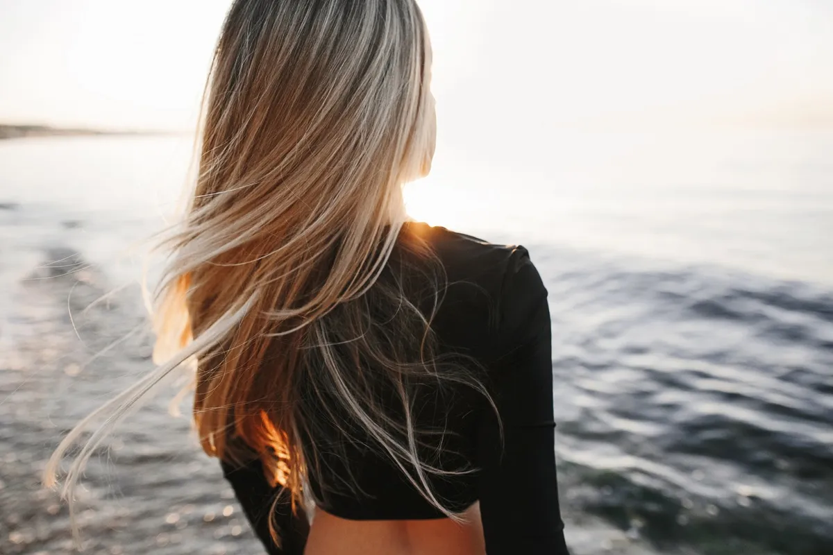 Donna di spalle con capelli biondi luminosi mossi dal vento, indossa una maglia nera e guarda il mare al tramonto.