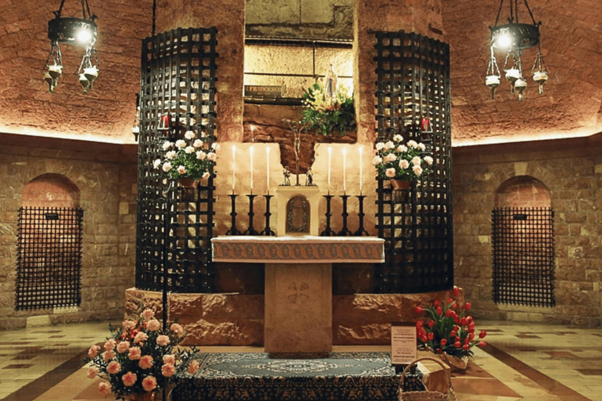 Altare principale nella cripta di San Francesco d’Assisi, con candele accese, fiori freschi e pareti in pietra illuminate da luci calde, creando un’atmosfera sacra e contemplativa.