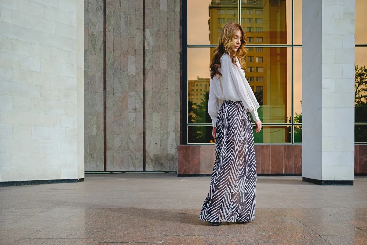 Donna indossa una gonna lunga estiva con stampa geometrica bianca e nera e camicia bianca leggera davanti a un edificio moderno con finestre a specchio.