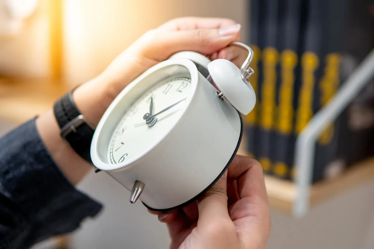 Mani di una persona che regolano un orologio analogico bianco, simbolo del cambio dell’ora legale o del risveglio mattutino.