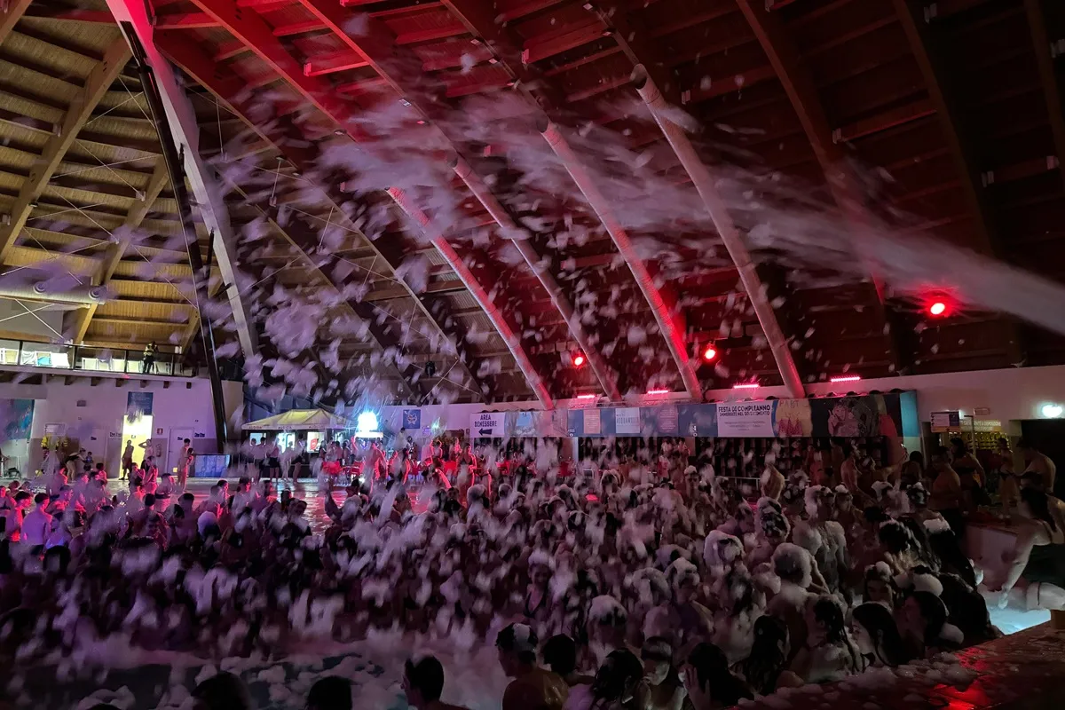 Persone che ballano immerse nella schiuma durante una festa notturna all’interno dell’Acquaworld, illuminate da luci rosse sotto la cupola del parco.