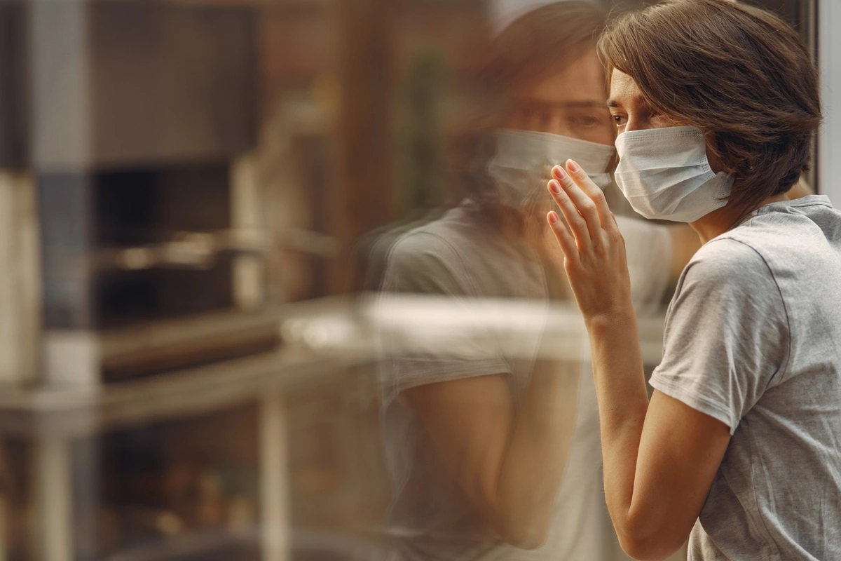 Donna con capelli corti e mascherina chirurgica guarda pensierosa fuori da una finestra, riflessa nel vetro, durante l’epidemia di influenza in Italia.