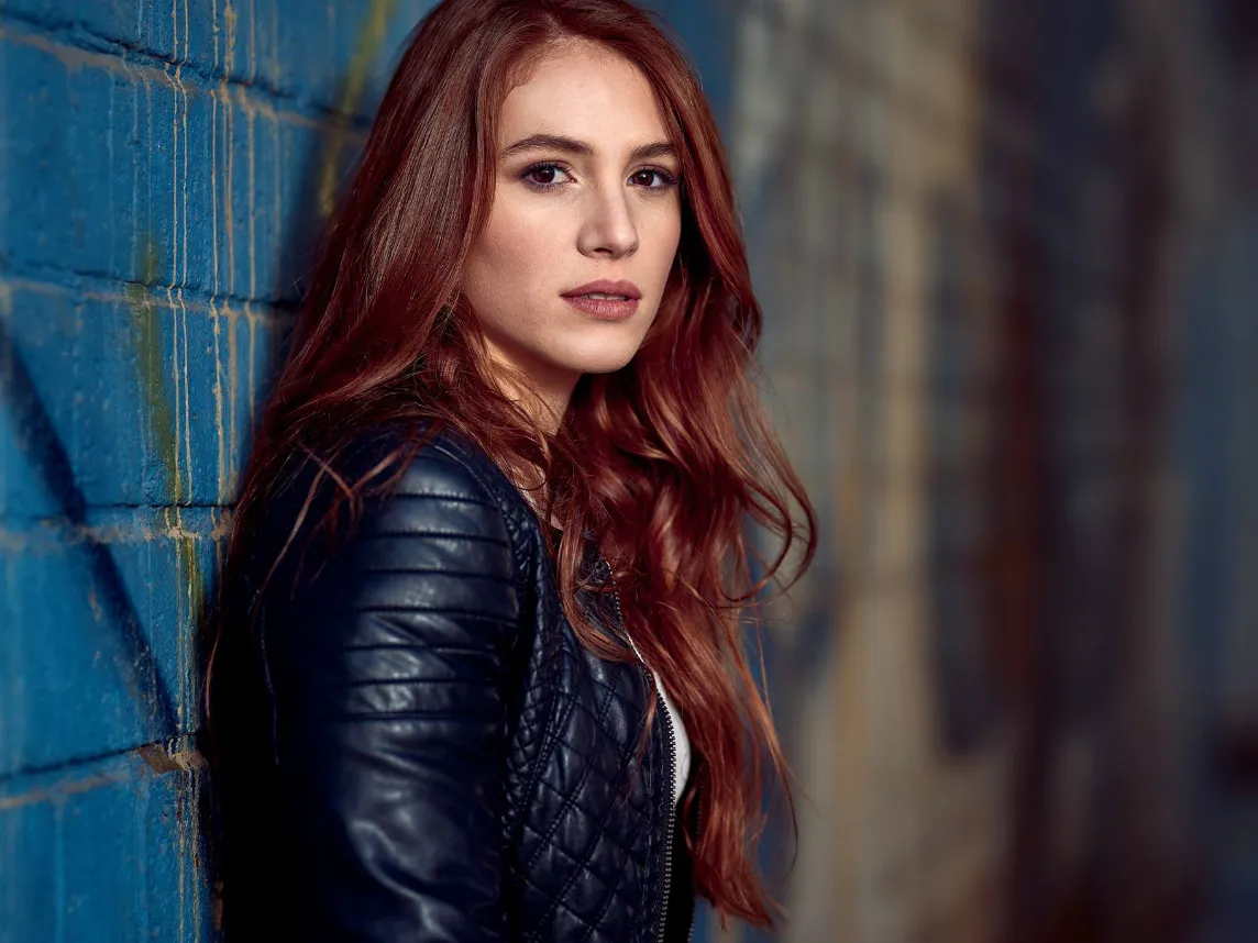 Giovane donna dai capelli rossi indossa una giacca di pelle nera e guarda verso la fotocamera, appoggiata a un muro blu con atmosfera urbana.