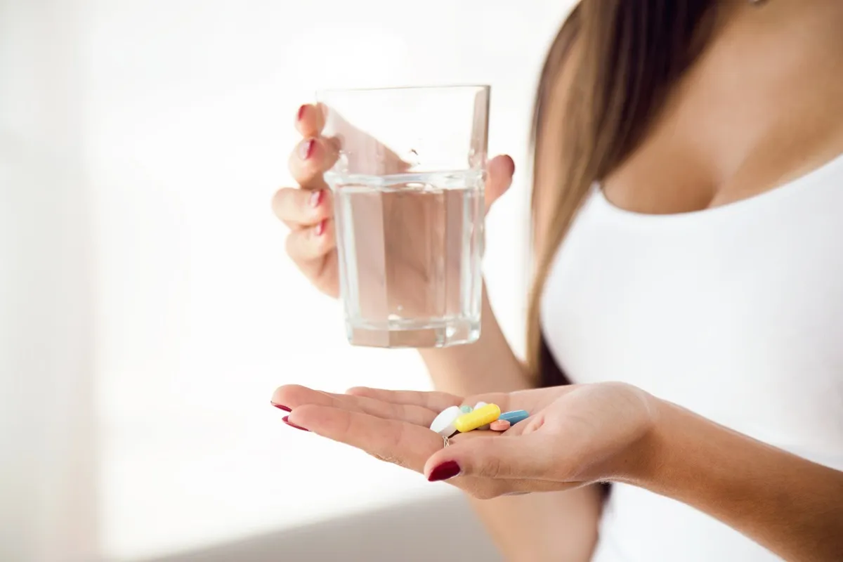 Donna con un bicchiere d’acqua in mano e diverse pillole colorate sull’altra mano, pronta ad assumere integratori alimentari.