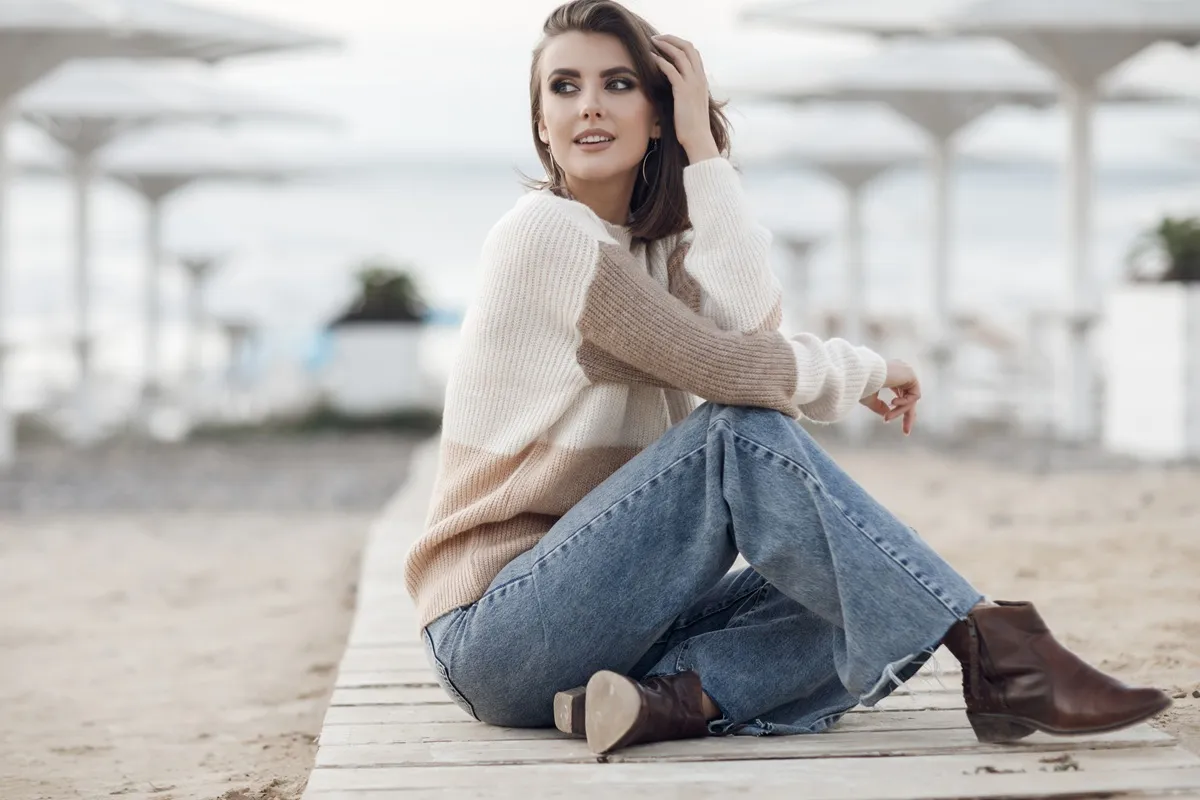 Donna elegante seduta sulla passerella di legno in spiaggia, indossa jeans palazzo, maglione a righe beige e stivaletti marroni.