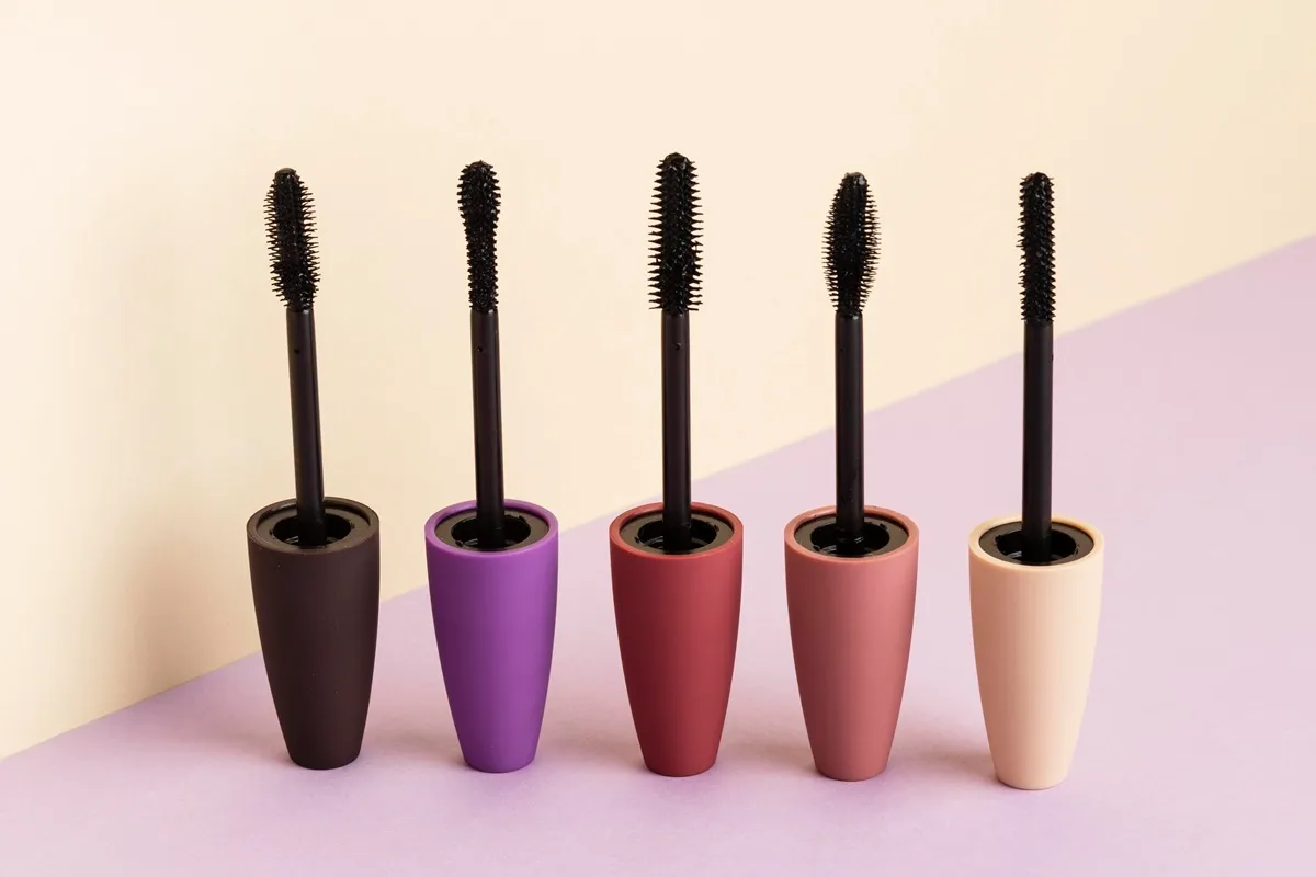 Cinque mascara aperti con scovolini neri e contenitori colorati disposti su fondo pastello.
