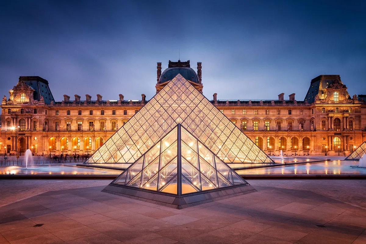 Vista serale del Museo del Louvre a Parigi con la piramide di vetro illuminata e il palazzo storico sullo sfondo.