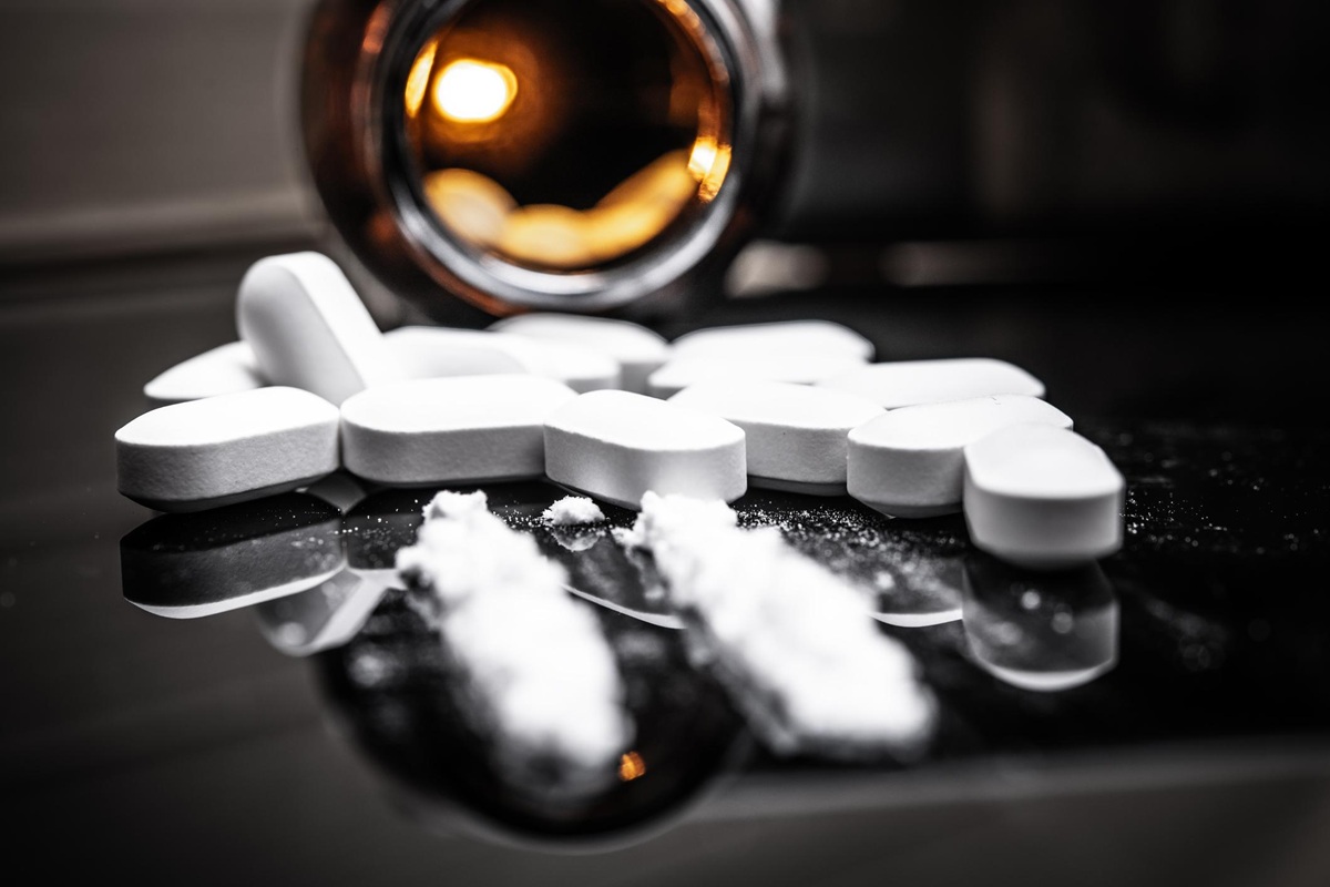 Compresse bianche sparse su una superficie nera accanto a un barattolo aperto, con polvere bianca in primo piano, simbolo di abuso di farmaci o sostanze.