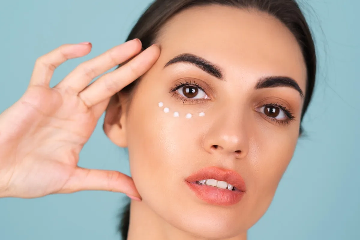 Donna applica crema contorno occhi con piccoli punti bianchi sotto l’occhio, mostrando la tecnica corretta di applicazione per idratare e ridurre le occhiaie.