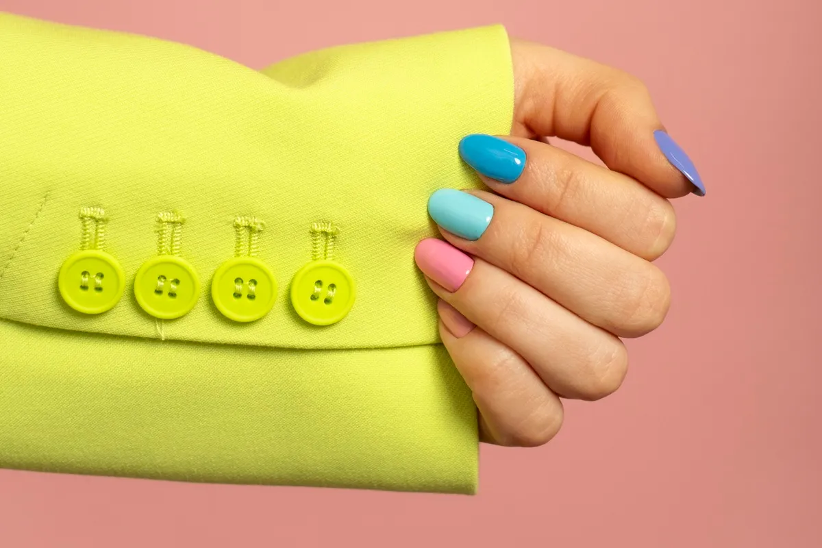 Mano con unghie colorate in tonalità pastello: blu, azzurro, rosa e lilla, su sfondo rosa e giacca gialla con bottoni decorativi.