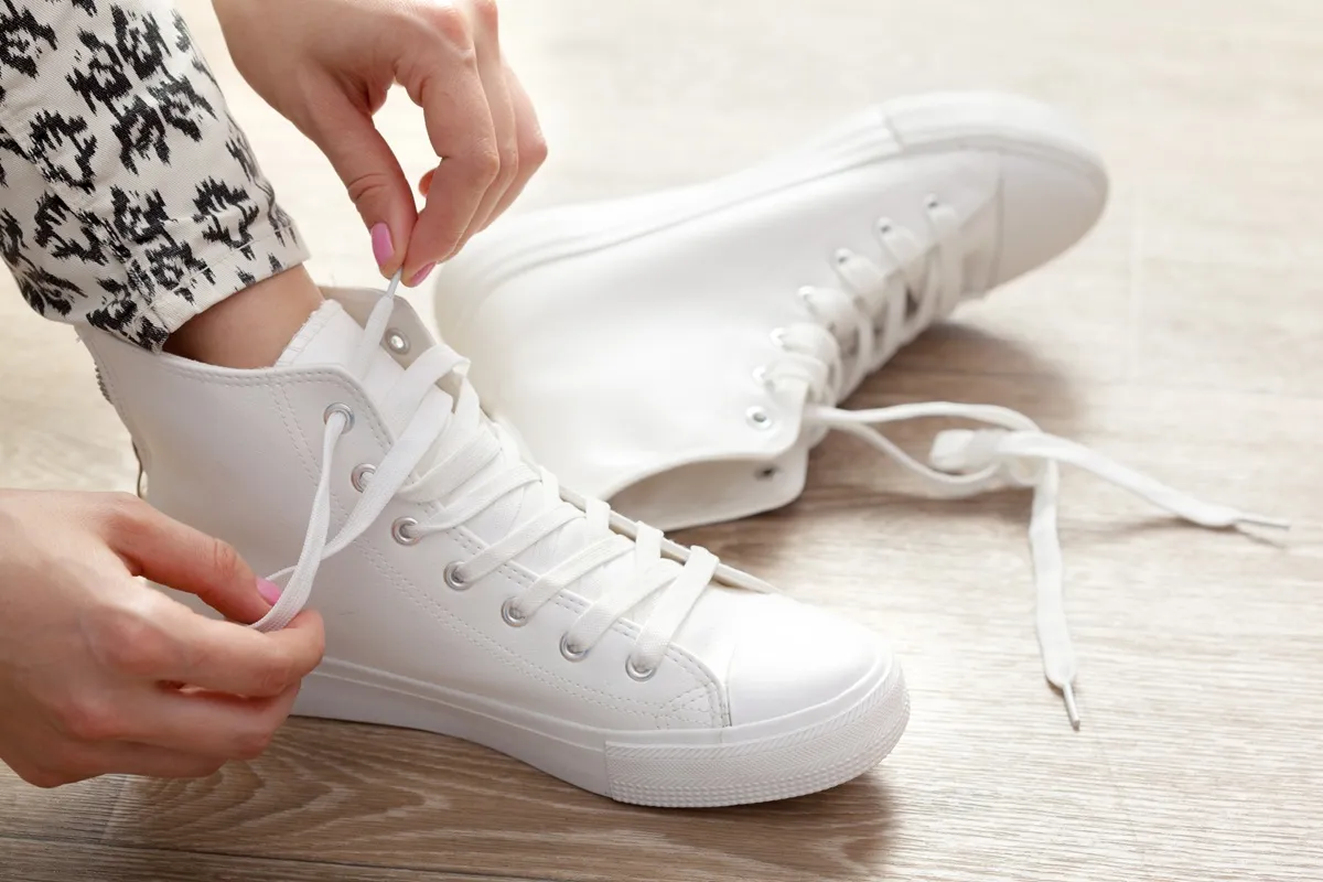 Donna allaccia sneakers bianche alte su pavimento in legno chiaro.