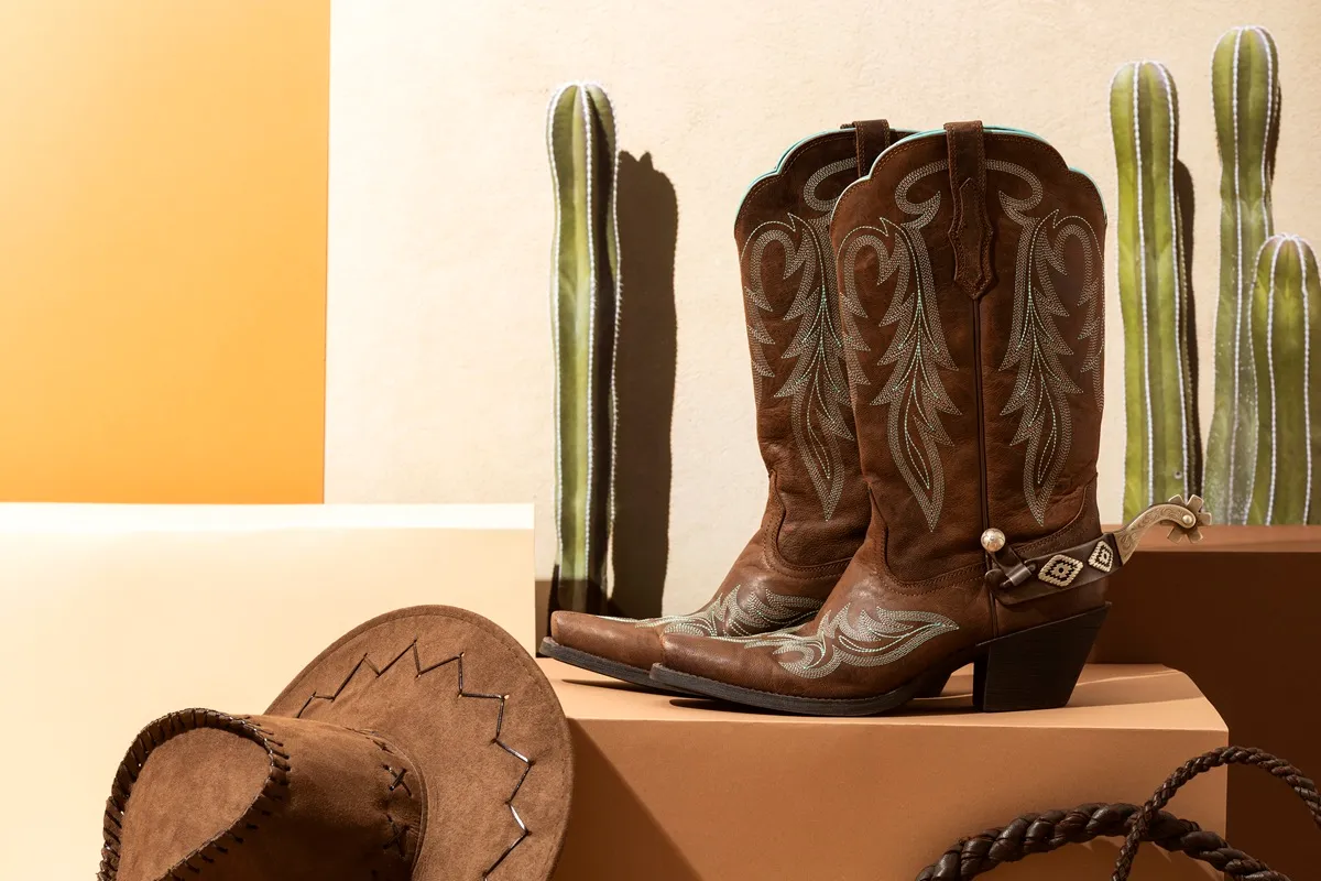 Stivali texani da donna in pelle marrone con dettagli ricamati, accanto a un cappello da cowboy e cactus decorativi