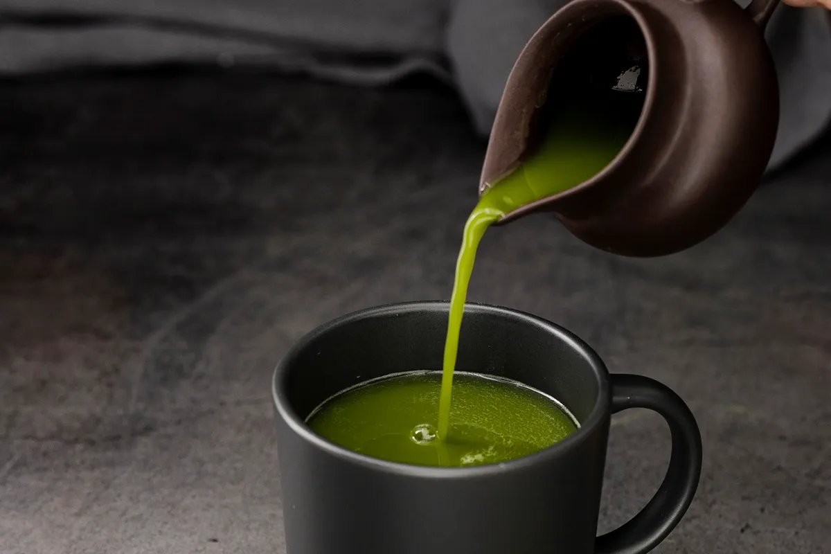 Tè matcha verde brillante versato da una teiera marrone in una tazza nera su sfondo scuro.