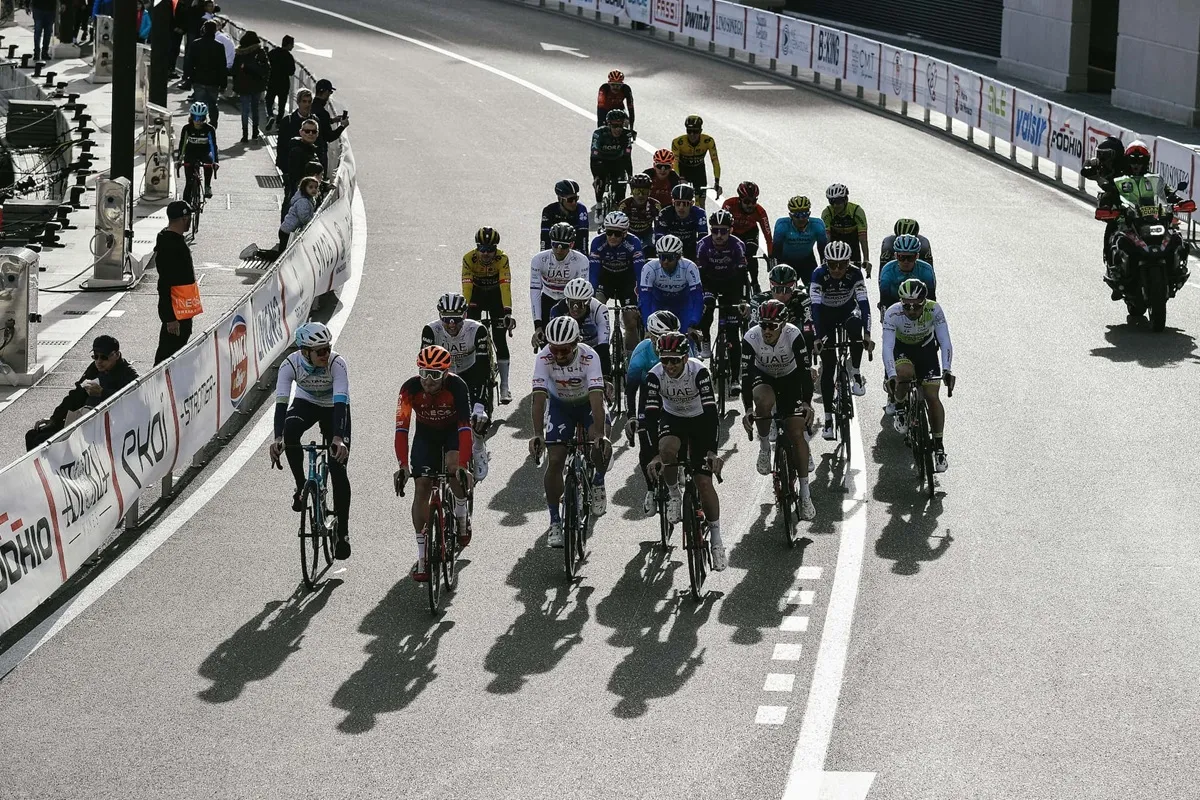Gruppo di ciclisti professionisti che percorre una strada cittadina durante una gara, sotto la luce del sole, seguiti da moto di supporto e osservati dal pubblico ai lati della carreggiata.