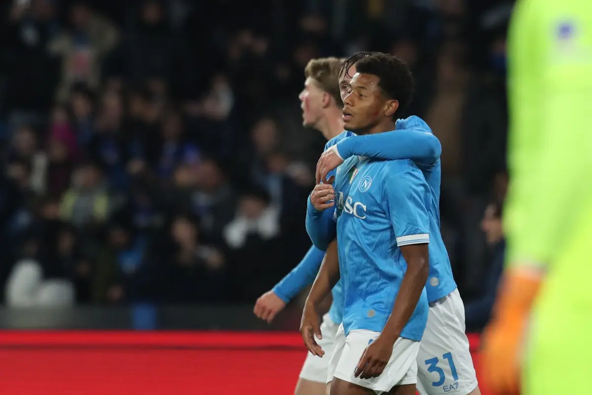 Calciatori del Napoli in maglia azzurra si abbracciano in campo durante una partita, con i tifosi sfocati sullo sfondo.