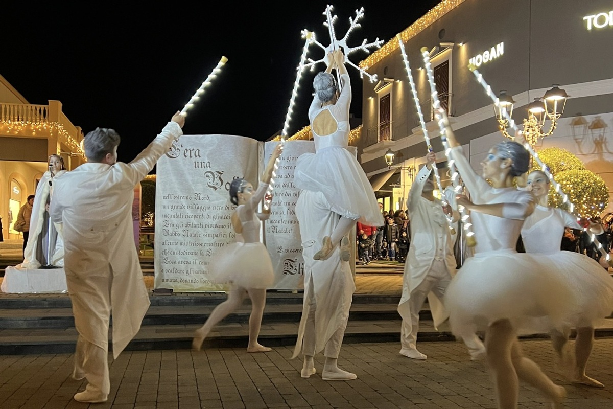 Danzatori in costumi bianchi si esibiscono nello spettacolo “Ice Dreams” al Sicily Outlet Village, circondati da luci natalizie e scenografie incantate, davanti a un grande libro decorativo.