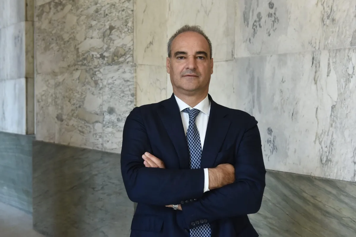 Ritratto di Alberto Bertone, fondatore di Acqua Sant’Anna, in posa con le braccia incrociate davanti a una parete di marmo, indossando un elegante abito blu e una cravatta decorata, simbolo di determinazione e leadership imprenditoriale.