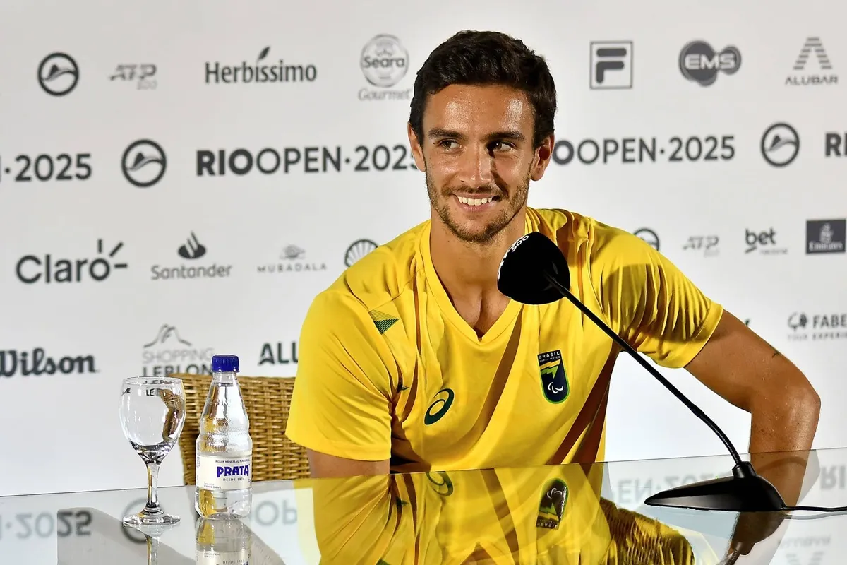 Lorenzo Musetti sorride durante una conferenza stampa al Rio Open 2025, indossando la maglia gialla della nazionale italiana di tennis, con microfono e bottiglia d’acqua davanti a sé.