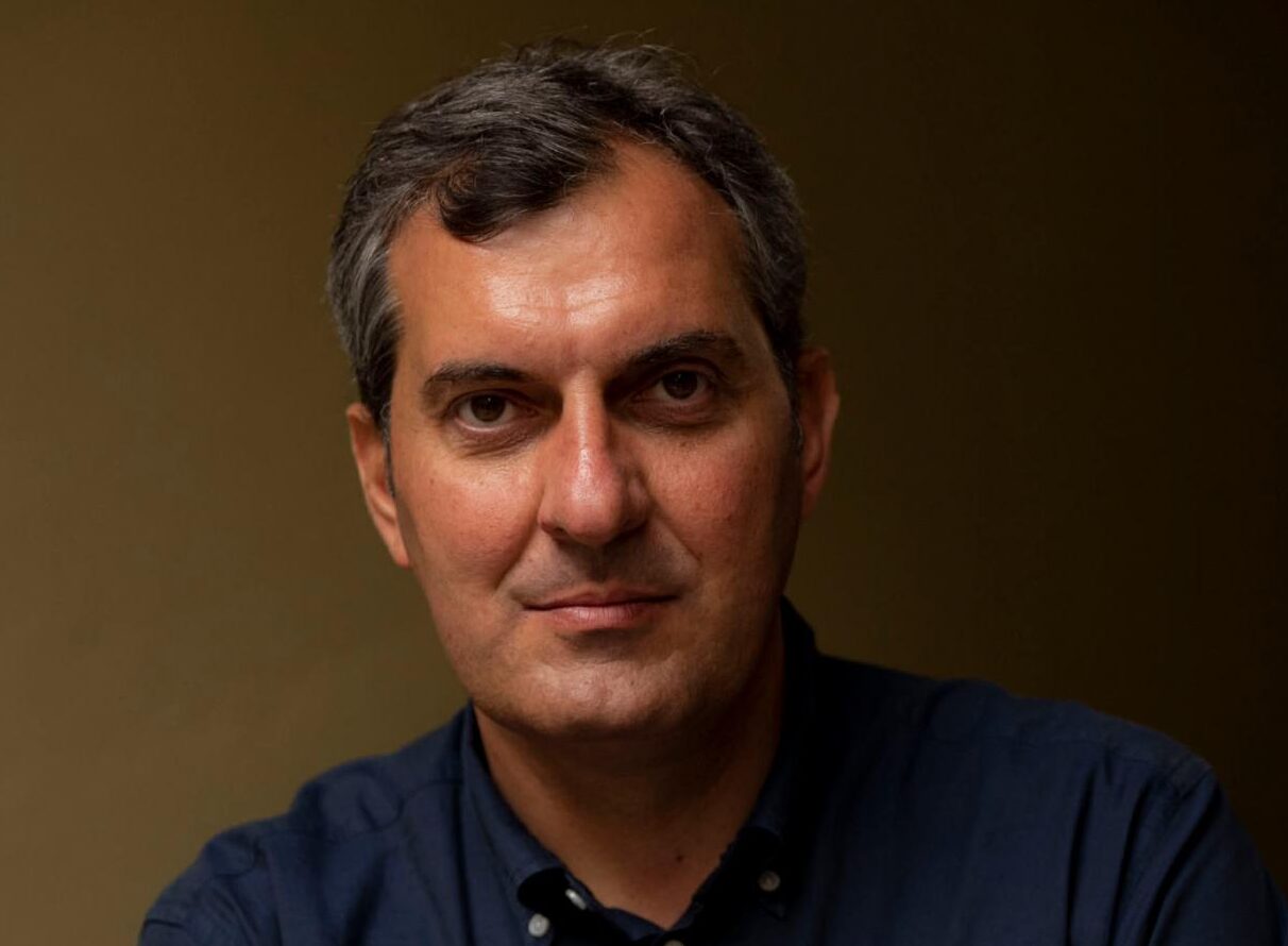 Primo piano di un uomo maturo con capelli brizzolati, espressione seria e sfondo neutro.