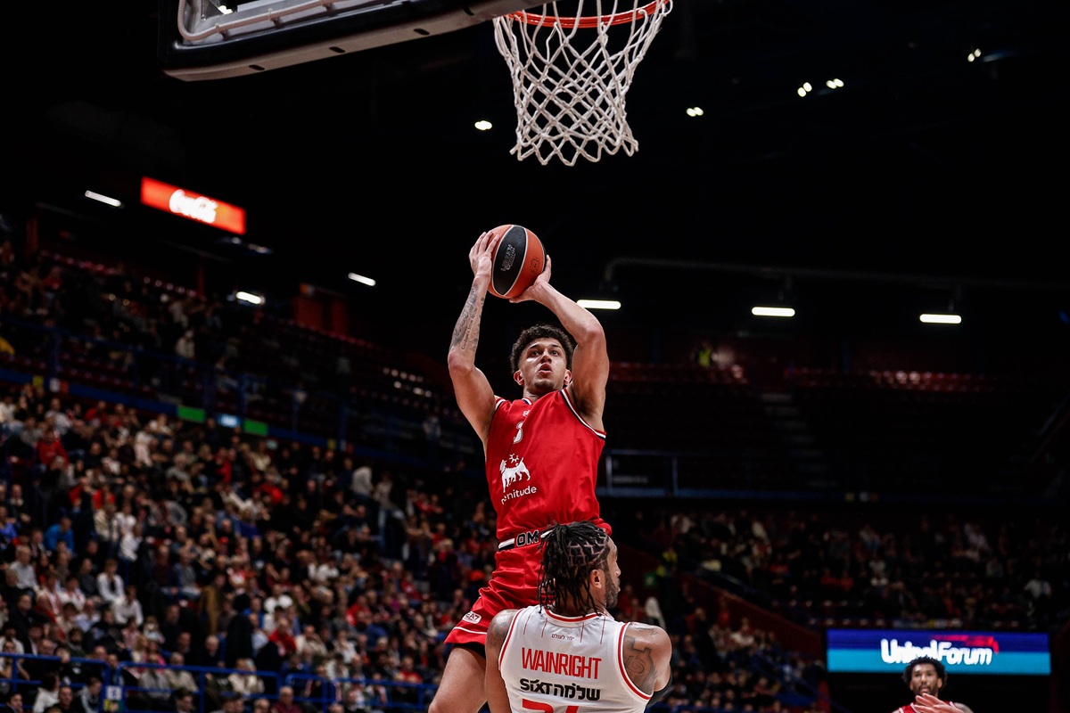Giocatore dell’Olimpia Milano in maglia rossa salta verso il canestro con la palla in mano durante una partita di basket Eurolega, mentre un avversario tenta di difendere.
