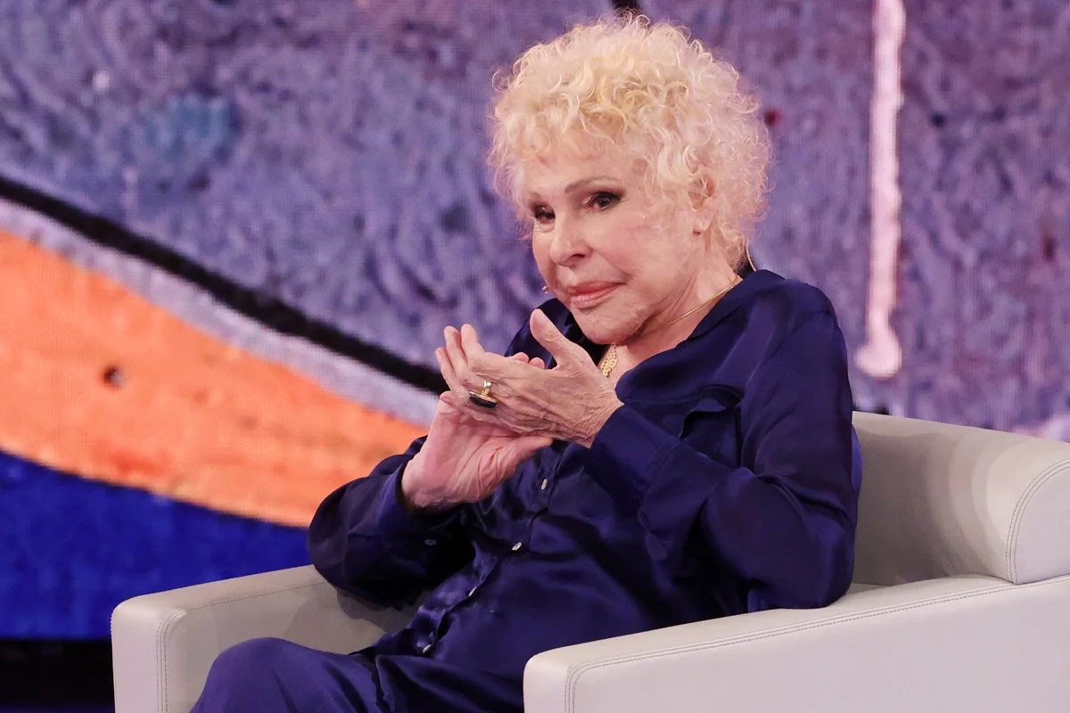 Ornella Vanoni seduta su una poltrona durante un programma televisivo, con capelli biondi ricci e abbigliamento blu scuro, guardando verso la camera in un’espressione serena.
