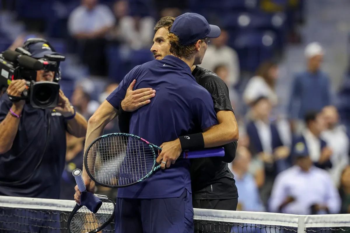 Jannik Sinner e Lorenzo Musetti si abbracciano a fine partita sul campo da tennis, mostrando rispetto e sportività, mentre un cameraman riprende la scena agli US Open.