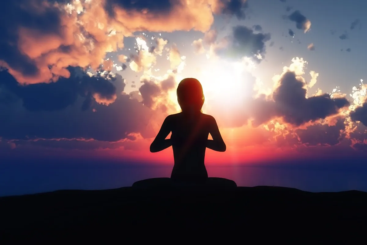Persona in meditazione al tramonto con il sole che filtra tra le nuvole, creando un’atmosfera di pace e spiritualità.