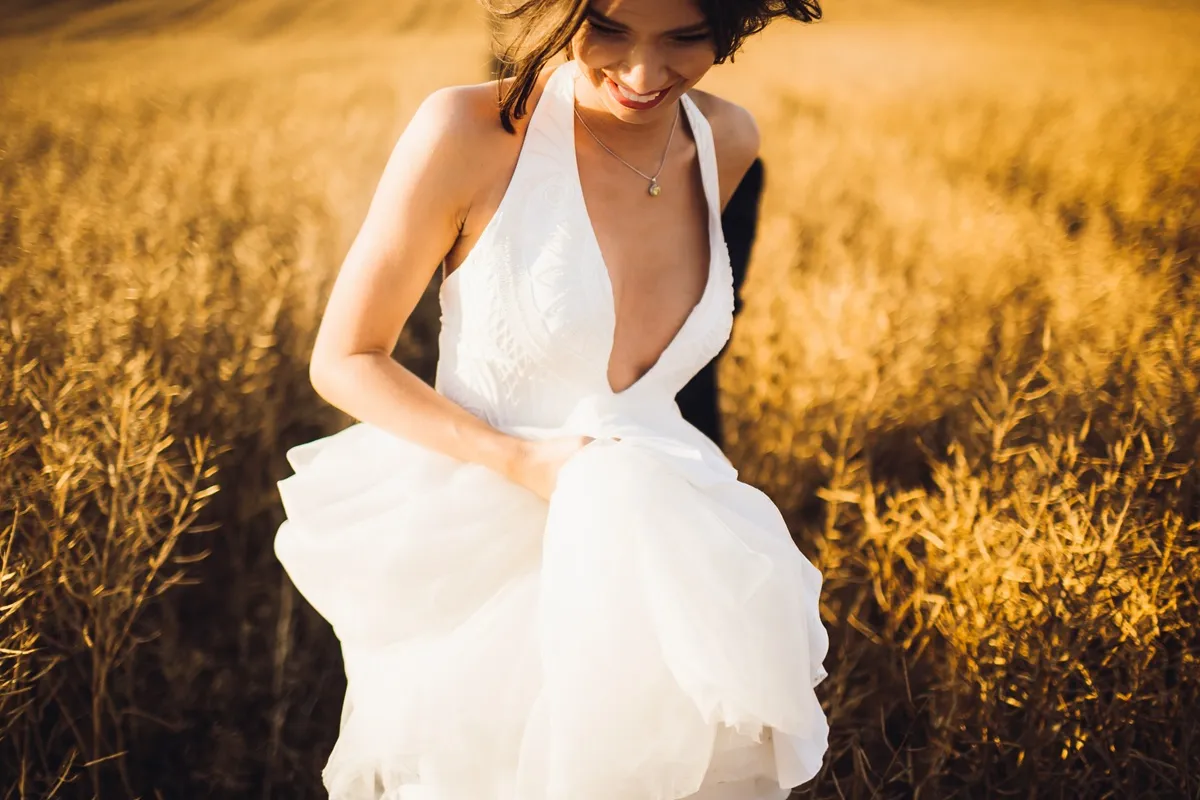Donna sorridente con abito da sposa bianco in stile boho chic cammina in un campo dorato illuminato dal sole al tramonto.