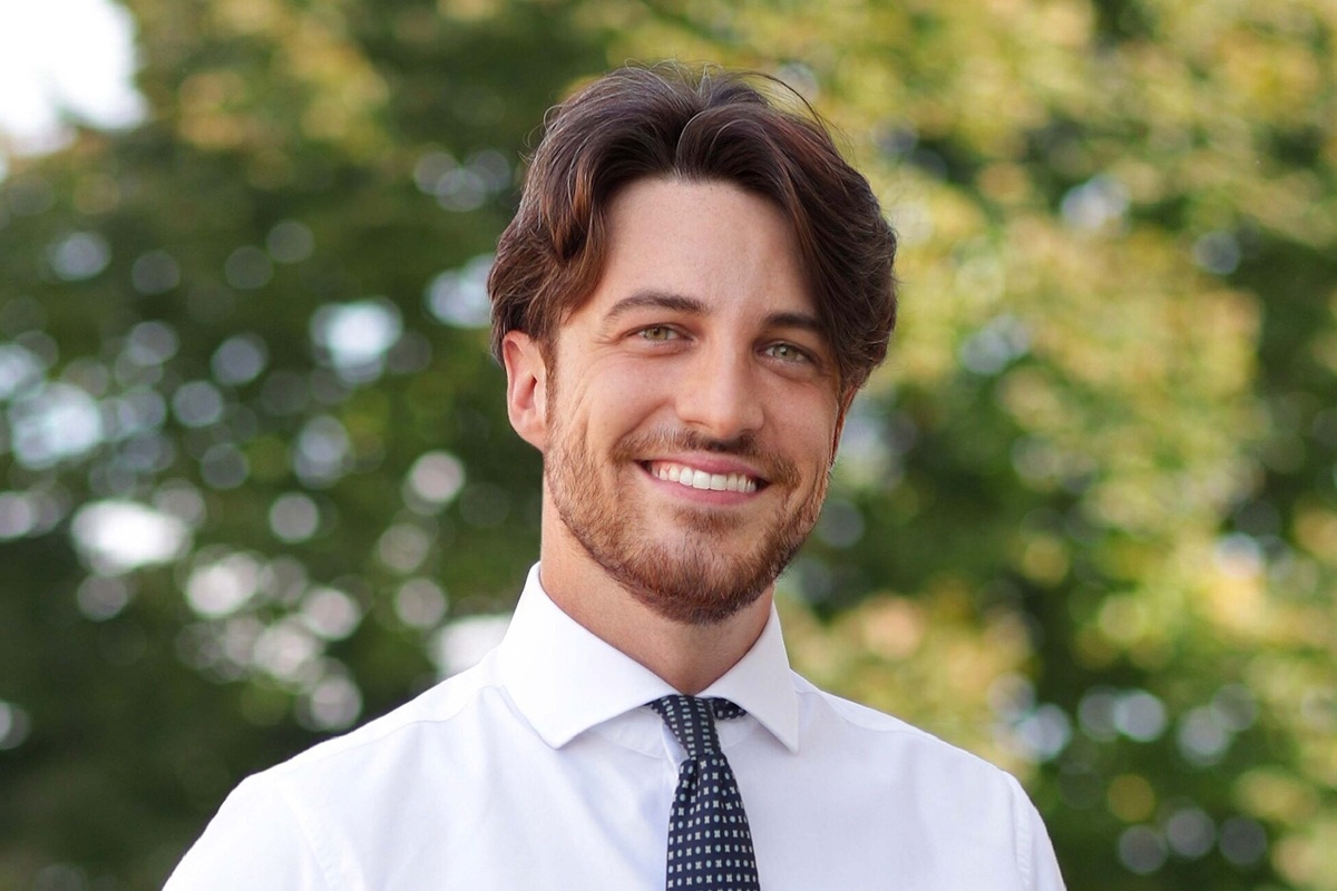 Giovane uomo sorridente con barba curata, camicia bianca e cravatta scura, fotografato all’aperto con sfondo verde sfocato.