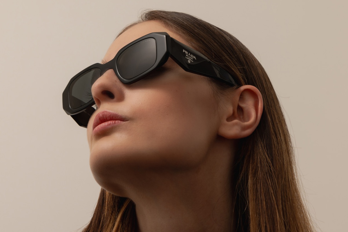 Donna con occhiali da sole Prada neri dal design geometrico, posa guardando verso l’alto su sfondo neutro, trasmettendo uno stile moderno e deciso.