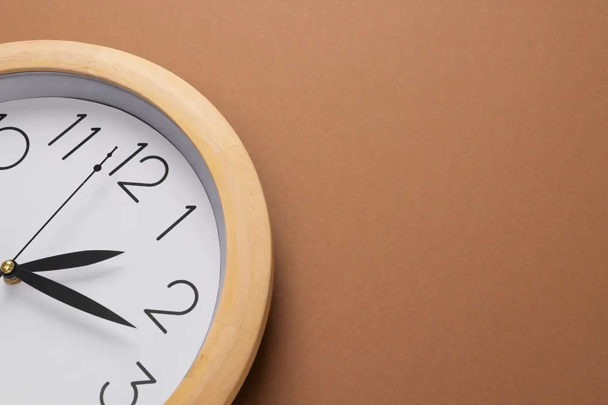 Orologio da parete con cornice in legno chiaro su sfondo marrone minimalista.