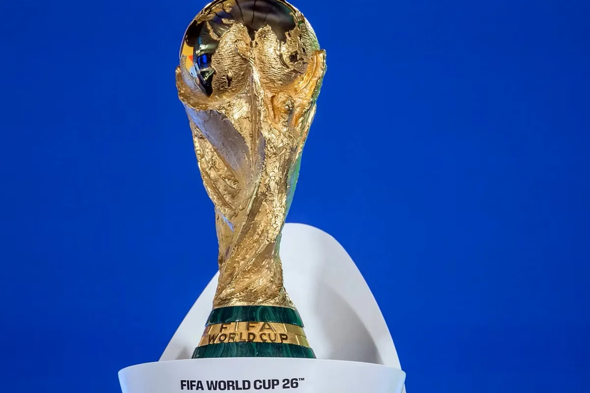 Trofeo dorato della Coppa del Mondo FIFA 2026 su sfondo blu, con base verde e dettagli metallici.