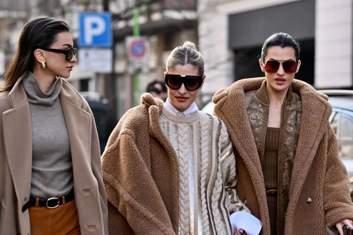 Tre donne camminano per le strade di Milano indossando cappotti oversize in tonalità neutre, maglioni a trecce e occhiali da sole scuri, esprimendo un look elegante e moderno tipico dello street style milanese invernale.