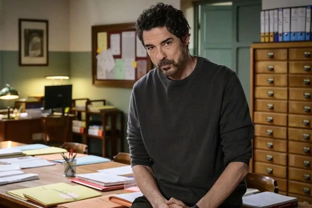 Uomo seduto su una scrivania piena di fascicoli in un ufficio scolastico, con armadi e bacheca alle sue spalle.