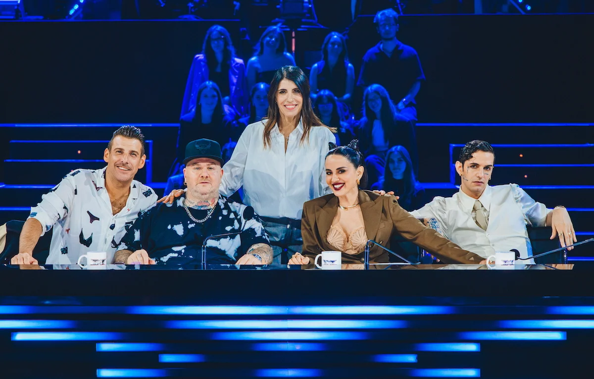 Foto della giuria di X Factor 2025 composta da cinque giudici seduti al tavolo, sorridenti, con pubblico illuminato sullo sfondo.
