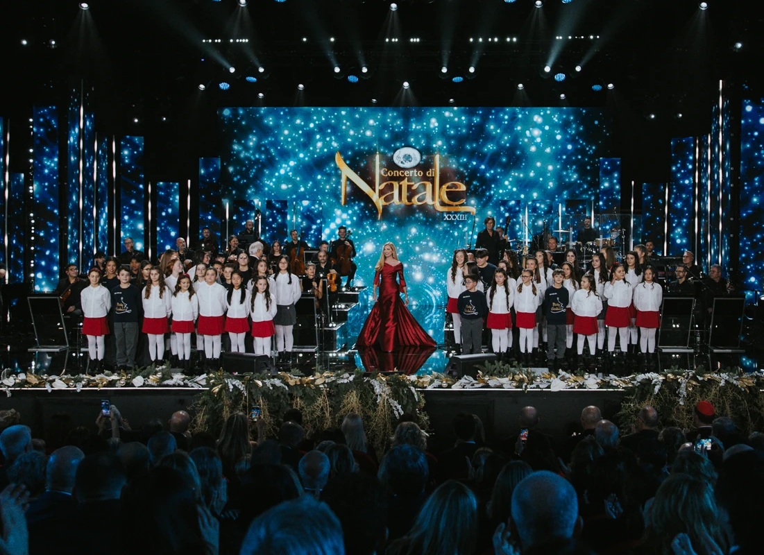 Concerto di Natale con orchestra sinfonica, coro di bambini e presentatrice in abito rosso su un grande palco illuminato