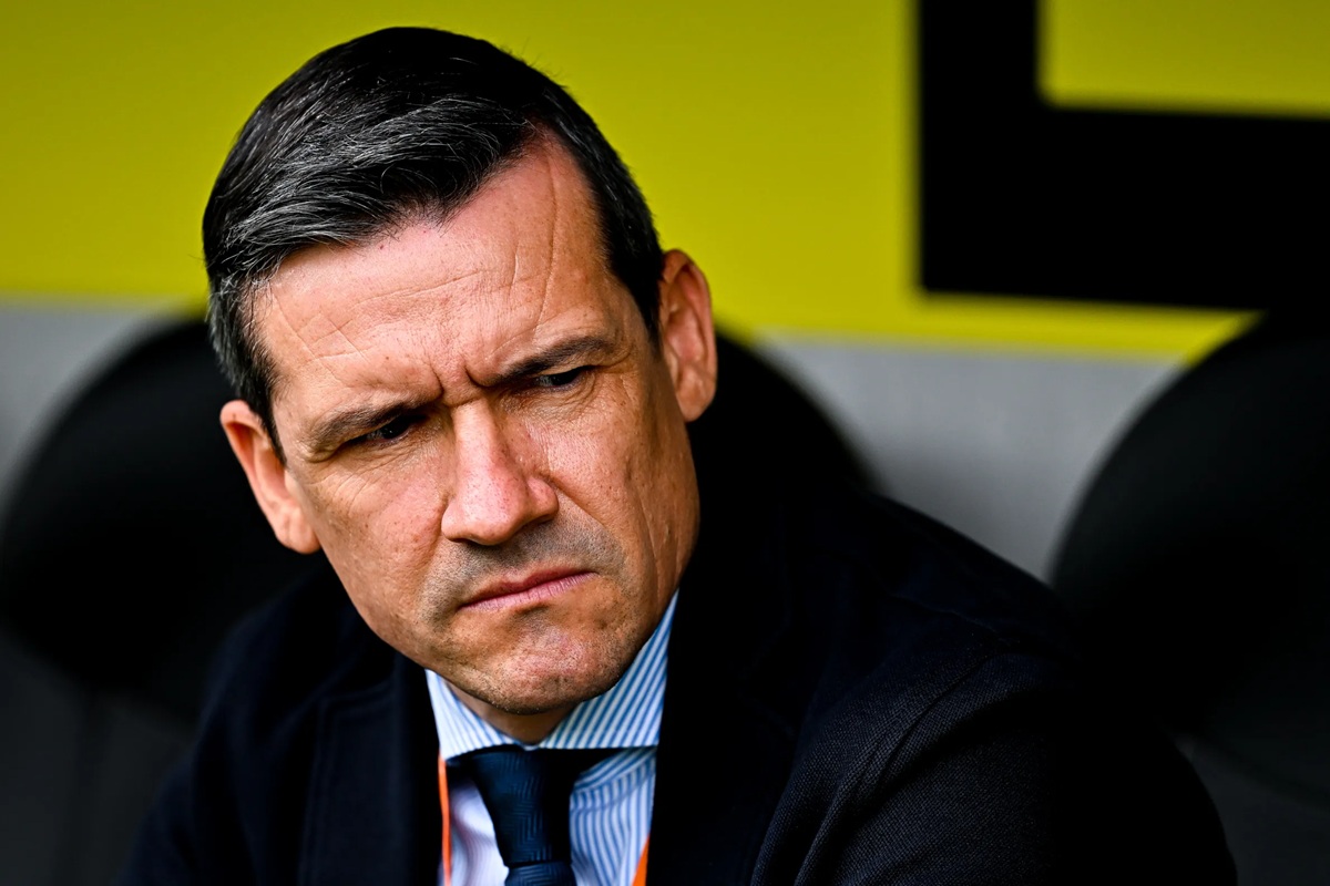 Dirigente sportivo in giacca e cravatta seduto in panchina, con espressione seria e concentrata durante una partita di calcio.