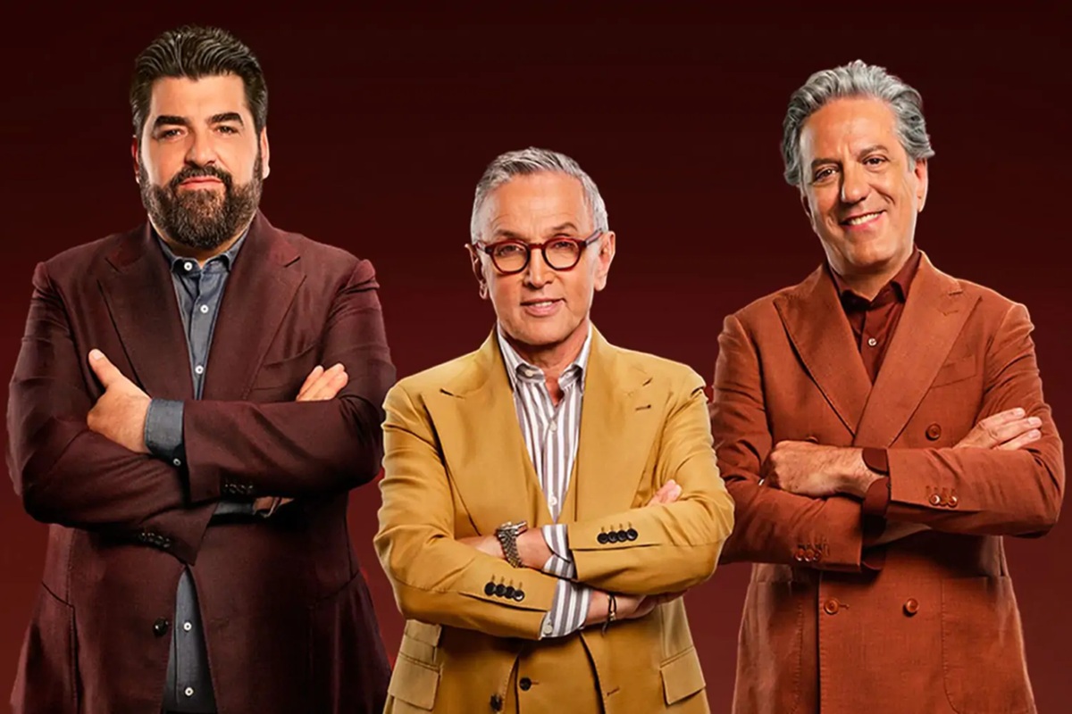 Tre giudici di MasterChef Italia in posa con le braccia incrociate, vestiti con completi eleganti su sfondo rosso.