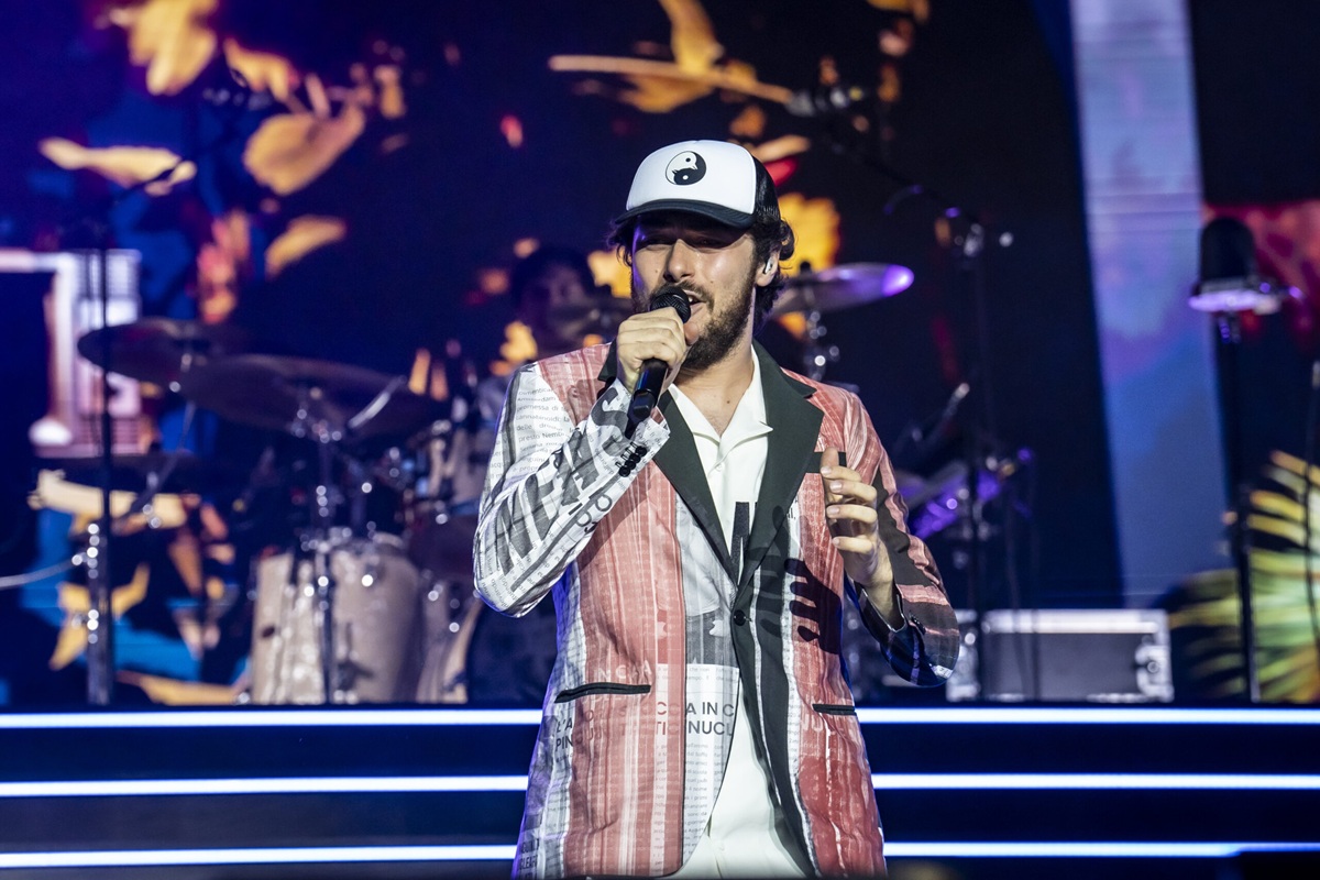 Cantante sul palco durante un concerto, indossa un completo colorato e un cappellino bianco mentre canta al microfono con una band sullo sfondo.
