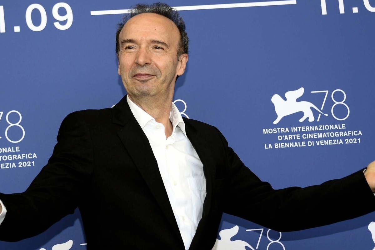 Uomo in giacca nera e camicia bianca posa sorridendo davanti al pannello blu della Mostra di Venezia